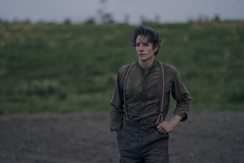 "Young man in old-fashioned clothes and suspenders stands in a field with a somber expression, overcast sky and open green landscape in the background."